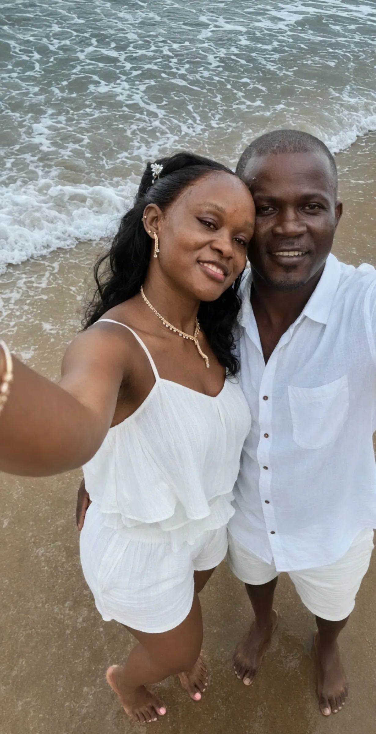 Richelle et Lionel, vêtus de blanc, posent pieds nus sur le sable humide au bord de l'océan.